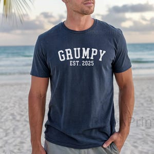 Peut inclure: Un t-shirt bleu marine avec un texte blanc qui dit "GRUMPY EST. 2025". La personne portant le t-shirt est debout sur une plage.
