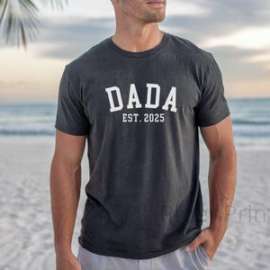 May include: A black t-shirt with white text that reads "DADA EST. 2025". The person wearing the shirt is standing on a beach.