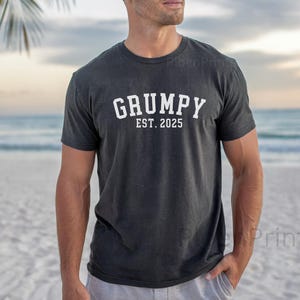 Op de afbeelding: Een donkergrijs T-shirt met witte tekst die "GRUMPY EST. 2025" zegt.