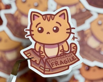 May include: A die-cut sticker featuring a cartoon orange tabby cat sitting inside a brown cardboard box. The box has the text EMOTIONALLY FRAGILE printed on the front flap. The cat has large dark eyes, pointed ears, and a light-colored belly.