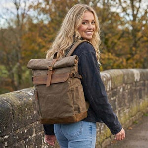 May include: A brown canvas backpack with leather detailing. The rucksack features a roll-top closure with a buckle and a front pocket. The bag is worn by a person wearing a black jacket and blue jeans.