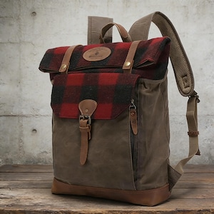 May include: A brown and red plaid backpack with leather straps and accents. The backpack has a flap closure and a side zipper pocket. The bag is made of canvas and leather, with a padded shoulder strap.