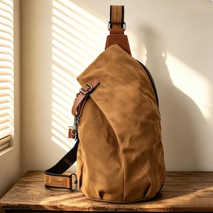 Op de afbeelding: Een bruine canvas sling bag met een leren schouderriem en accenten. De tas heeft een ritssluiting en een metalen sluiting. De tas staat op een houten oppervlak.