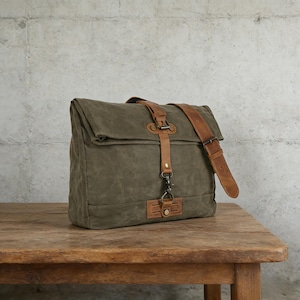 May include: Olive green canvas messenger bag with brown leather accents. Features a flap closure, adjustable leather shoulder strap, and leather details on the front. The bag is sitting on a wooden table.
