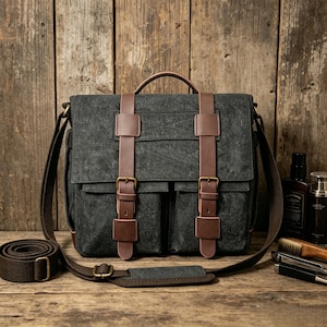 May include: A dark gray canvas messenger bag with brown leather accents. The bag features a top handle, adjustable shoulder strap, and two front pockets secured by leather straps and buckles. The bag is on a wooden surface.