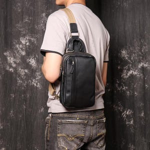 May include: A black leather sling bag with a vertical design, worn over the shoulder. The bag has a zippered main compartment and a smaller front pocket. The strap is tan and adjustable. The bag is worn by a person wearing a gray t-shirt and jeans.