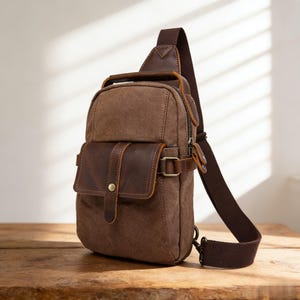 May include: A brown canvas and leather sling bag with a single shoulder strap. The bag features a front pocket with a leather flap and a metal buckle closure. The bag has a top handle and a zippered main compartment. The bag is sitting on a wooden surface.
