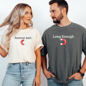 Puede incluir: Dos personas con camisetas a juego. Una camiseta es de color crema con el texto "Forever Isn't" y un gráfico de imán. La otra camiseta es gris con el texto "Long Enough" y un gráfico de imán. Ambas camisetas tienen mangas enrolladas.