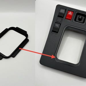 Op de afbeelding: Zwart auto-interieurpaneel met diverse knoppen en een rode waarschuwingsknipperlichtknop. Er wordt ook een apart zwart frame getoond. Het paneel heeft knoppen voor raambediening en andere functies. De items staan op een witte achtergrond.