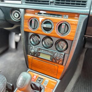 Puede incluir: Vista interior del salpicadero de un coche clásico. El salpicadero presenta molduras de madera, una radio y varios indicadores. También se ven la palanca de cambios y las rejillas de ventilación. El interior del coche es negro y gris.