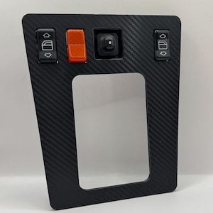 Op de afbeelding: Een zwart bedieningspaneel voor auto's met een koolstofvezelpatroon, voorzien van twee zwarte raamschakelaars met witte iconen, een rechthoekige oranje waarschuwingslichtschakelaar en een centrale zwarte instelknop voor zijspiegels.