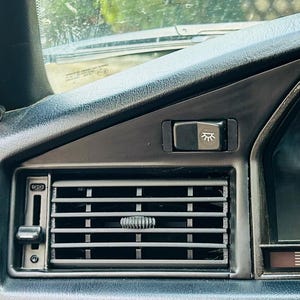 Könnte beinhalten: Nahaufnahme eines schwarzen Armaturenbretts im Auto mit einem Lüftungsgitter mit verstellbaren Lamellen und einem Schalter. Das Armaturenbrett umfasst auch einen Lichtschalter und einen Teil des Tachometers.