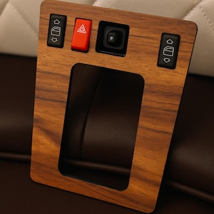 Op de afbeelding: Een houten interieur trimstuk voor auto's met raam- en waarschuwingslichtbediening. De trim heeft een houtnerf afwerking en heeft zwarte knoppen met witte pictogrammen en een rode waarschuwingslichtknop. De trim heeft een rechthoekige opening.