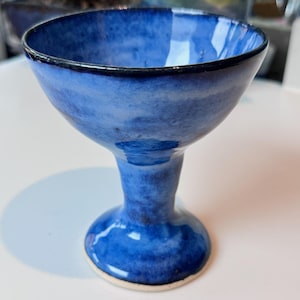 May include: A handmade, blue ceramic goblet with a wide bowl and a slender stem. The goblet has a dark blue rim and a white base. The blue glaze has a marbled effect, creating a unique, artistic look. This piece is perfect for serving beverages.
