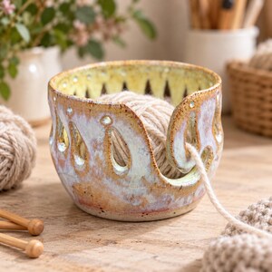 May include: A ceramic yarn bowl with a speckled, multi-coloured glaze in shades of yellow, blue and brown. The bowl features a series of teardrop-shaped cutouts around the rim and a spiral opening on the side for feeding yarn through while knitting.