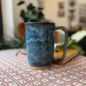 May include: A handmade ceramic mug with a blue glaze and a brown rim. The mug has a large handle and a cylindrical shape. The mug is sitting on a pink and white patterned tablecloth. The mug is ideal for coffee or tea.