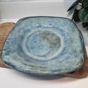 Può includere: Un piatto quadrato in ceramica con smalto blu e bianco. Il piatto ha una forma leggermente irregolare e un bordo rialzato. Il piatto è appoggiato su un tagliere di legno. Il piatto misura circa 20 cm di larghezza.