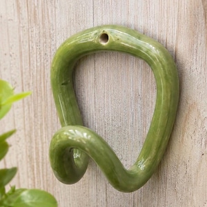 May include: A glossy, olive green ceramic wall hook with a fluid, organic, and twisted shape. The hook features a single circular mounting hole at the top centre, designed for attachment to a flat surface, showcasing a smooth, reflective finish.