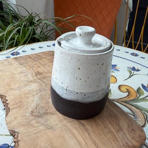 May include: A handmade ceramic jar with a lid. The jar is cylindrical, with a speckled white upper section, a gray band, and a dark brown base. The lid has a small knob. The jar sits on a wooden cutting board.