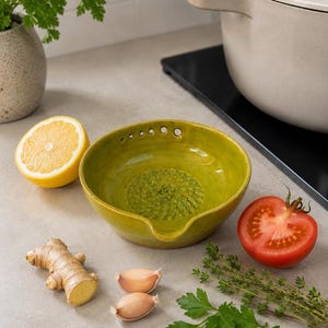 May include: A lime green ceramic garlic grater bowl with a textured interior, next to a halved lemon, a tomato, ginger, garlic cloves, and herbs. A beige cooking pot is in the background.