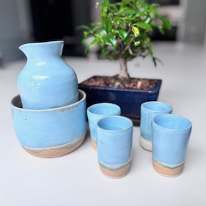 May include: A light blue ceramic sake set, including a pitcher, bowl, and four small cups. The set has a rustic, handmade look with a light brown base. A small bonsai tree in a blue pot is in the background.