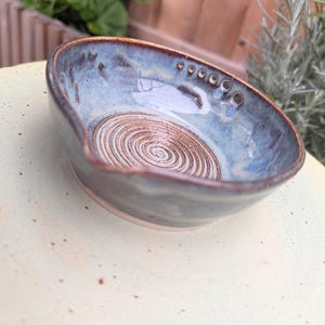 May include: A small, round ceramic spoon rest with a blue and brown glazed finish. The interior features a spiral pattern at the base and a series of small circular indentations along the upper rim. The edges are finished in a dark brown tone.