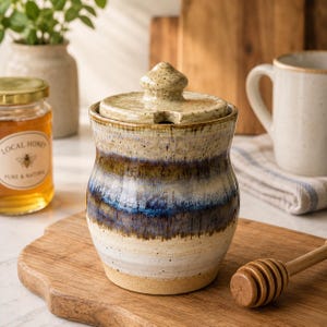May include: A ceramic honey pot with a lid, featuring a speckled cream base that transitions into horizontal bands of deep blue, brown, and tan glaze. The pot has a rounded body and a small knob on the lid, designed to hold a wooden honey dipper.