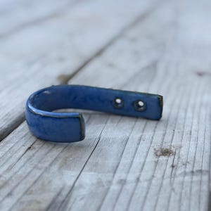 May include: A U-shaped ceramic wall hook featuring a glossy blue glaze with subtle tonal variations. The hook has a smooth, rounded finish and includes two pre-drilled circular holes on the flat mounting end for secure installation.