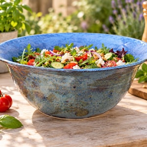 May include: A large, round, ceramic serving bowl with a wide, flared rim. The exterior features a mottled, textured finish in shades of deep blue, brown, and grey, while the interior rim is a vibrant, solid blue. The bowl is filled with a fresh salad.
