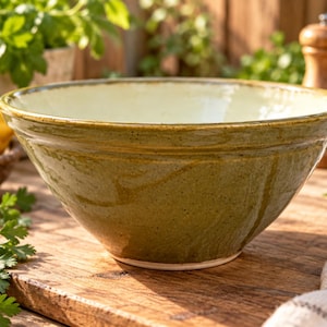 May include: A wide, round ceramic bowl with a glossy, mottled olive-green exterior and a smooth, cream-coloured interior. The bowl features a thick, rounded rim and a slightly tapered base, showcasing a rustic, hand-crafted appearance.