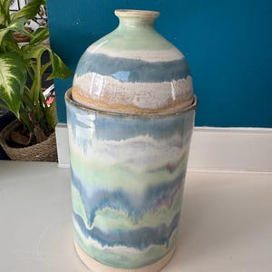 May include: A ceramic jar with a lid. The jar is cylindrical with a rounded top and lid. The jar is painted with horizontal bands of blue, green, and white. The lid is a matching color scheme. The jar sits on a white surface.