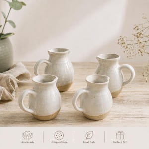 Può includere: Quattro piccole brocche in ceramica con smalto bianco sporco lucido e basi in argilla beige a vista. Ogni brocca presenta un corpo arrotondato, un manico curvo e un bordo svasato, disposte su una superficie in legno chiaro.