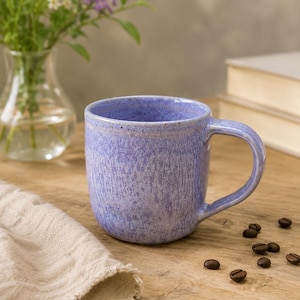 Peut inclure: Un mug en céramique avec un émail moucheté lavande et violet clair. Le mug présente un corps arrondi, une anse incurvée sur le côté droit et une finition légèrement texturée avec des variations subtiles d'intensité de couleur.