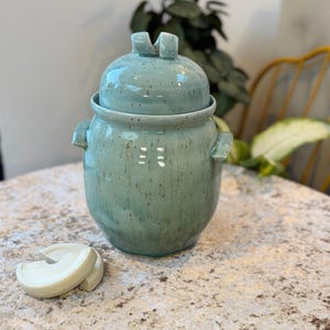 Könnte beinhalten: Ein türkisfarbener Keramiktopf mit Deckel, kleinen Griffen und einer gesprenkelten Oberfläche. Der Deckel hat ein einzigartiges Griffdesign. Ein separates, cremefarbenes Keramikstück liegt vor dem Topf. Der Topf steht auf einer gemusterten Oberfläche.