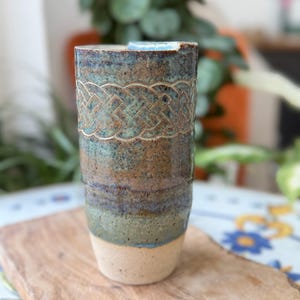 May include: A handmade ceramic tumbler with a Celtic knot design. The tumbler has a textured surface with a gradient of colours, including brown, teal, and blue. The bottom is a light beige colour. The tumbler is approximately 15 cm tall.