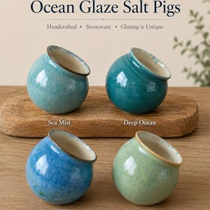 Può includere: Quattro saliere in gres di forma rotonda con aperture inclinate, etichettate come Ocean Glaze Salt Pigs. I colori sono Sea Mist, Deep Ocean, Blue Tide e Sea Green. Ogni pezzo presenta una finitura lucida con un bordo distinto e varie tonalità di blu e verde.