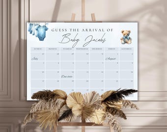 Op de afbeelding: Een blauw en wit baby shower-spel met een kalender waarop gasten de aankomstdatum van Baby Jacobs kunnen raden. De kalender omvat de maanden juli en augustus. Het spel bevat een blauwe teddybeer met een strikje en een waslijn met babykleding.