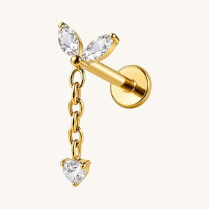 Puede incluir: Un pendiente de botón con parte posterior plana en tono dorado, con dos piedras transparentes de corte marquesa en forma de hoja, unidas a una cadena dorada corta que termina en una pequeña piedra transparente de corte triangular.