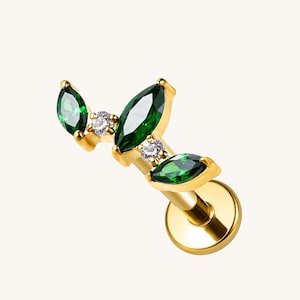 May include: A gold-tone flat back stud earring featuring a curved branch design. The piece is adorned with three marquise-cut dark green stones and two small round clear stones set in a polished gold metal frame with a circular base plate.