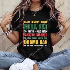 May include: Black t-shirt with text in green, red, and yellow. The text reads "Black History Month," "Rosa Sat," "So Martin Could Walk," "Martin Walked," "So That Obama Could Run," and "Obama Ran."