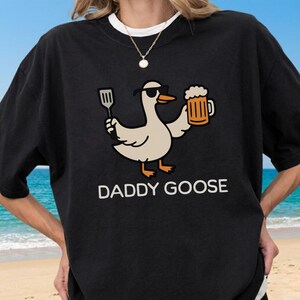 May include: Black t-shirt featuring a cartoon goose wearing sunglasses and holding a spatula and a beer mug. The text "DADDY GOOSE" is printed in white below the graphic. The shirt has a crew neck and short sleeves.