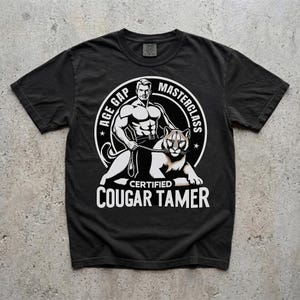 May include: Black t-shirt with a white graphic. The graphic features a muscular man holding a leash attached to a cougar. Text on the shirt reads "AGE GAP MASTERCLASS CERTIFIED COUGAR TAMER".