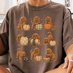 Op de afbeelding: Een bruin T-shirt met korte mouwen en een grafische print van negen bruine puppy's die met verschillende pompoenen met patronen spelen. De pompoenen hebben diverse ontwerpen, waaronder bloemen-, ruit-, stippen- en stermotieven, geaccentueerd door kleine oranje blaadjes.