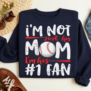 May include: Navy blue sweatshirt with the text "I'M NOT just his MOM I'm his #1 FAN" in white and red. A baseball is featured in the center of the design. The sweatshirt is a crew neck style.