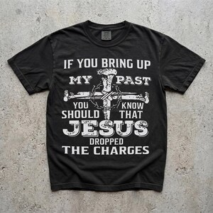 May include: Black t-shirt with white text: "If you bring up my past, you should know that Jesus dropped the charges." A cross and crown of thorns graphic is centered above the text. The shirt is made of a soft material.