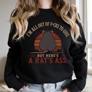 Peut inclure: Un sweat-shirt noir à col rond avec un motif représentant un rat gris vu de dos, centré sur un cercle aux couleurs du coucher de soleil. Le texte indique : I'M ALL OUT OF F*CKS TO GIVE BUT HERE'S A RAT'S ASS. Le design utilise des lettres beige et rouge.
