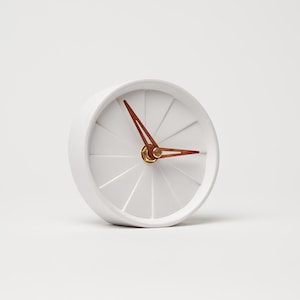 May include: White round clock with a white face and copper-colored hands. The clock has a simple design with a clean, minimalist aesthetic.