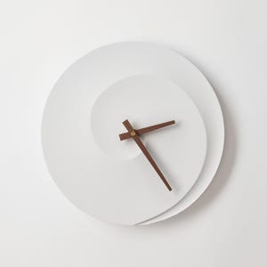 Könnte beinhalten: Weiße Wanduhr mit einem Spiraldesign. Die Uhr hat Holzzeiger und eine minimalistische Ästhetik. Das Zifferblatt hat ein einzigartiges, geschichtetes Aussehen und erzeugt einen modernen und künstlerischen Look. Der Durchmesser beträgt ca. 30 cm.