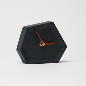 May include: A black hexagonal clock with gold hands and a wooden second hand. The clock is sitting on a white surface.