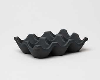 May include: A black egg carton made of a textured material. The carton has six individual compartments for holding eggs.